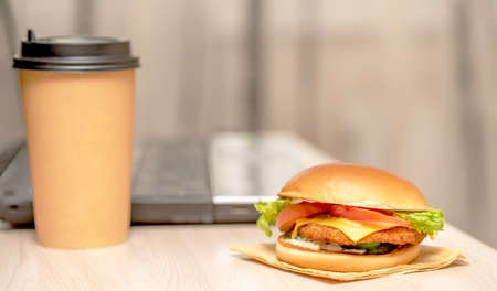 A hamburger close-up lies on the table. In the background there is a computer and a disposable glass of coffee.の写真素材