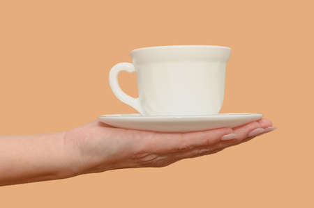 A female hand holds a white cup and saucer. minimalism. close-up. On a beige background.の写真素材