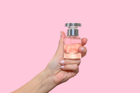 A woman's hand holds a glass bottle of perfume. On a pink background. close-up.
