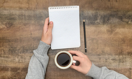 Male hands hold a notebook and a cup of coffee on a brown table. view from above.