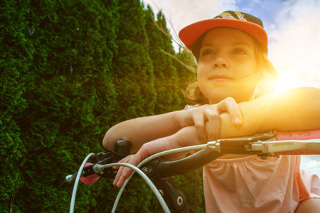 Young girl cyclist enjoy the beautiful sunrise on summer forest trail. Close-up portrait of a happy little girl in a helmet on a bicycle. High quality photoの写真素材
