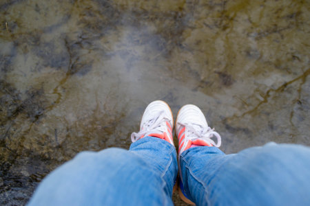 Selfie of woman's legs in sneakers and blue jeans standing in water.