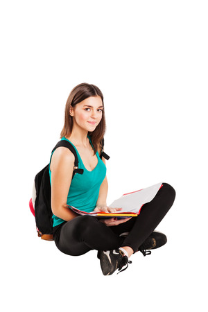 Young teen female student sitting on floor with backpack reading a book, isolated on whiteの写真素材