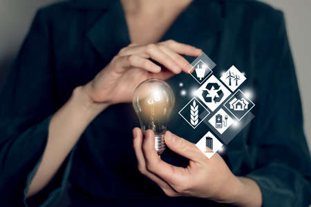 Energy innovation light bulb with future industry of power generation icons graphic interface in woman`s hands. Concept of sustainable development and alternative energy. Ecology concept.の写真素材