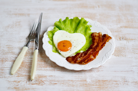Tasty Fried Egg in the Shape of a Heart Served on a White Plate with Bacon Tomato Salad Leaves Pepper Valentine Dayの写真素材