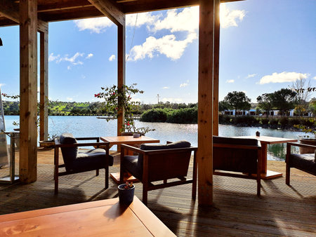 A peaceful lakeside scene captured from a sunlit wooden terrace, featuring cozy outdoor seating, blue skies and calm sparkling waterの写真素材