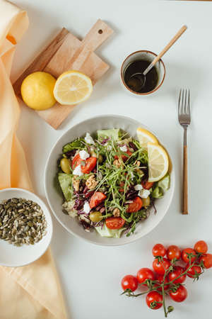 Healthy fresh salad of vegetables, cucumber, tomato, spinach, arugula in a plate on a white table, pumpkin and sunflower seeds with yellow lemon on wodden board. Food background, recipe. From above flat layの写真素材