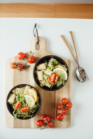 Two black bowl of rigatoni pasta with arugula, green peas, basil and cherry tomato served on wooden board. Italian food concept, flat layの写真素材