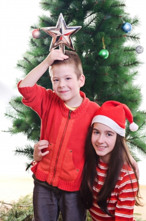 Young boy holding star on top of his head and sister wearing santa hat in front of christmas tree, portrait, looking at camera, vertical shotの写真素材