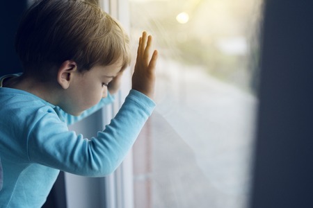 Little boy at home looking trough the window waiting for his mother to come back homeの写真素材