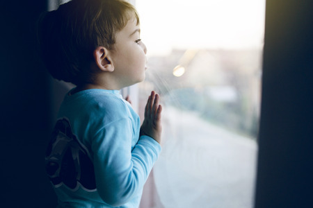 Little boy at home looking trough the window waiting for his mother to come back homeの写真素材