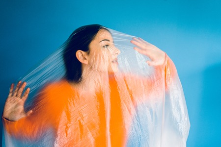 Portrait of young woman in orange in front of blue wall covered with nylon plastic bag, antipollution pollution save the planet oceans ecology conceptの写真素材
