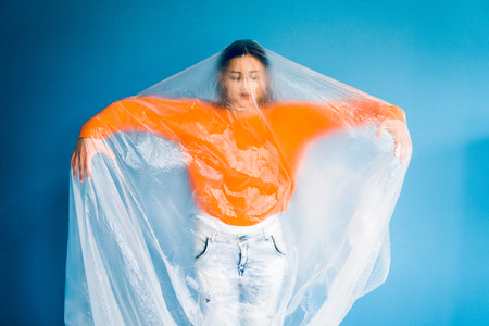Portrait of young woman in orange in front of blue wall covered with nylon plastic bag,...