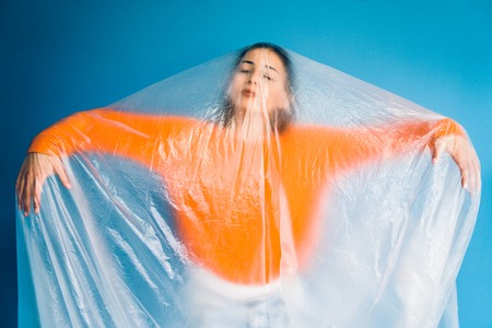 Portrait of young woman in orange in front of blue wall covered with nylon plastic bag, antipollution pollution save the planet oceans ecology conceptの写真素材