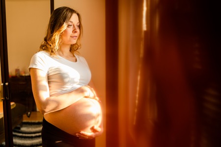 Young beautiful pregnant woman standing by the window looking holding her belly waiting for the babyの写真素材