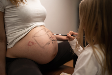 Little blonde girl painting on her mothers pregnant belly with water colorsの写真素材