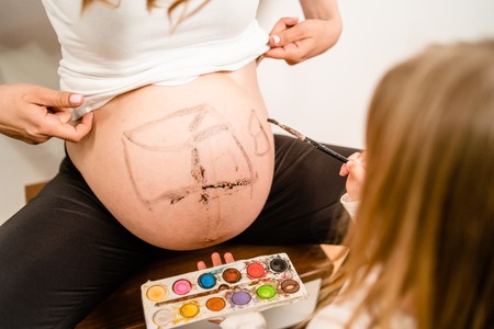 Little blonde girl painting little baby brother or sister on her mothers pregnant belly with water colors and paint brushの写真素材