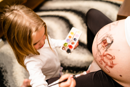 Little blonde girl painting little baby brother or sister on her mothers pregnant belly with water colors and paint brushの写真素材