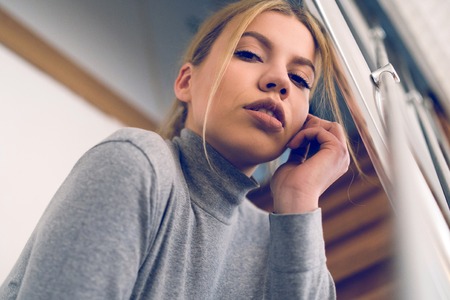 Portrait of beautiful young woman with blonde hair looking to the camera sitting on the stairs at homeの写真素材