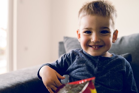 Portrait of a little small boy eating unhealthy snacks corn peanut flips at homeの写真素材
