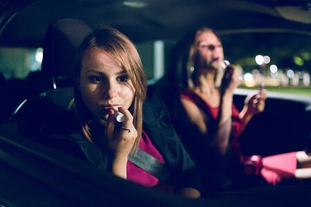 Two women mother and daughter friends putting make up repair lipstick in the car at night getting ready to go outの写真素材