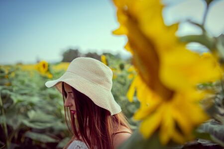 Portrait of young woman in white dress standing in the crops field of sunflowers in a sunny summer day wearing hat thinking emotionの写真素材