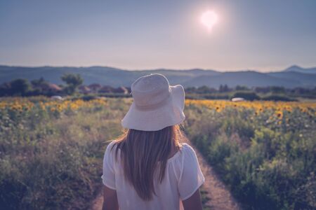 Portrait of young woman in white dress with hat standing in the dirt road in summer day walking away back viewの写真素材