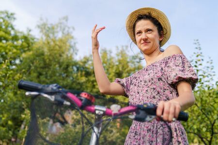 Young woman holding handlebar grip driving pushing bicycle in a summer sunny day wearing dress and hat in nature countryside outdoorの写真素材