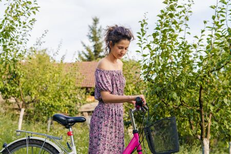 Young woman holding handlebar grip driving pushing bicycle in a summer sunny day wearing dress in nature countryside outdoorの写真素材