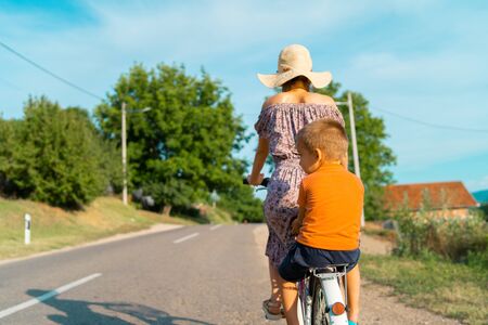 Young woman in a sunny summer day wearing dress riding a bike bicycle on the open road on the asphalt with her small boy kid child son in the back sittingの写真素材