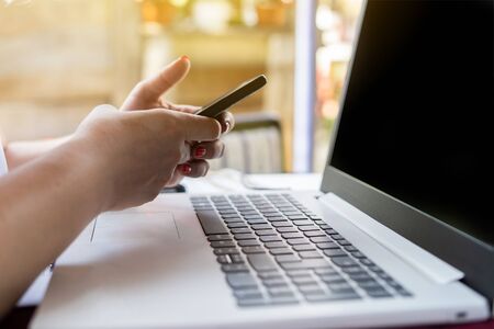 Close up on female woman hands holding smart mobile phone in garden above the laptop computer typing...