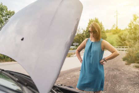 Young woman tourist driver standing by the road by the car with the hood open dead engine broken...