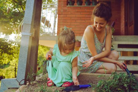 Young mother and daughter digging picking ground dirt land in their yard garden near the home house family planting flowers or seeds using gardening tools in summer dayの写真素材