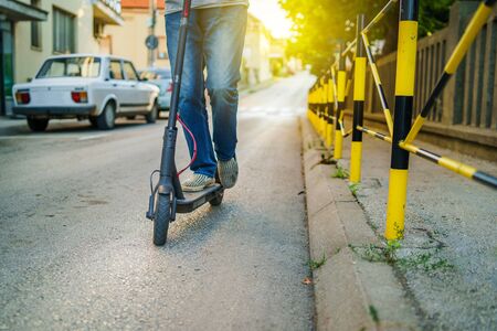 close up of senior man legs in jeans riding electric kick scooter on the street in summer evening day by the pavement fenceの写真素材