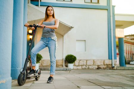 Young woman standing by electric kick scooter by the building in the town city waiting in summer day alternative transportの写真素材
