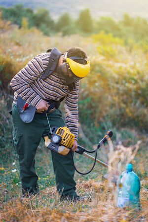 Young farmer gardener cutting grass weeds in the orchard plum plantation using trimmer petrol...
