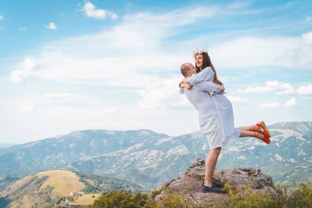 Young couple man and woman in love wearing white shirt and dress sitting hugging on the top of the mountain in summer spring or autumn dayの写真素材