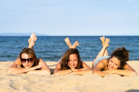 Three young women girl friends or sisters laying on the sand beach by the sea or ocean having fun in...