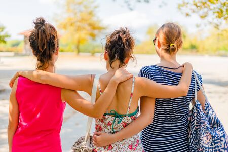 Vacation holiday back rear view of group three female woman girl friends hug hugging while walking in autumn or summer on the rest area by the road during travelの写真素材
