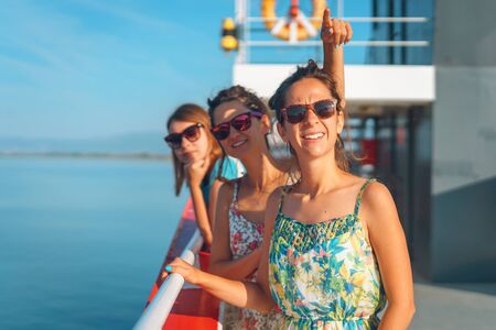 Three young women on the deck of the ship ferry boat talking pointing finger to the destination or sky showing smiling having fun on the vacation friends in sunny dayの写真素材