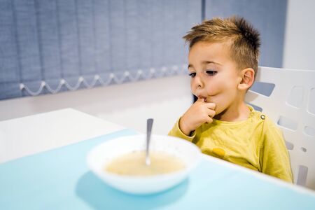 Portrait of small boy child eating soup meal or breakfast having lunch by the table at home with spoon white kindergarten finger in mouthの写真素材