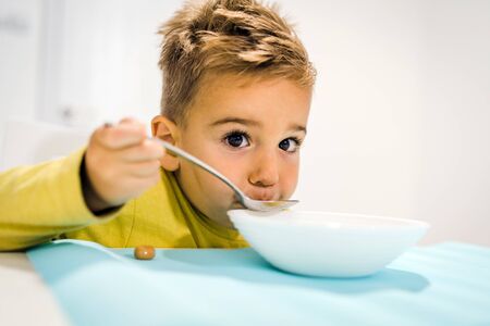 Portrait of small boy child eating soup meal or breakfast having lunch by the table at home with spoon white kindergartenの写真素材