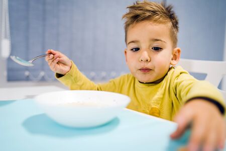 Portrait of small boy child eating soup meal or breakfast having lunch by the table at home with spoon white kindergartenの写真素材