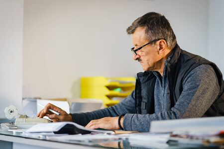 Senior man entrepreneur at his office store by the table accounting check bill calculation receipt making invoice for selling goods salesman at warehouse depot storehouseの写真素材