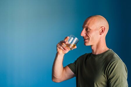Portrait of young handsome man with short hair holding a glass of whiskey or brandy alcohol drink standing in front of blue wall side view profileの写真素材