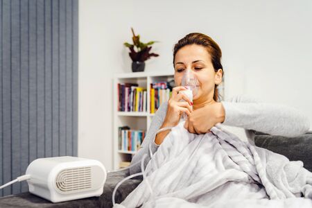 Woman lying covered with blanket using vapor steam inhaler nebulizer mask inhalation at home on the...