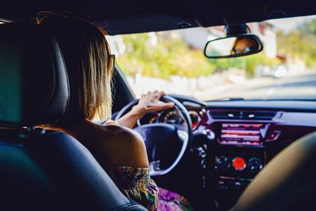 Woman driving a car wearing summer dress and sunglasses in a sunny autumn day back view
