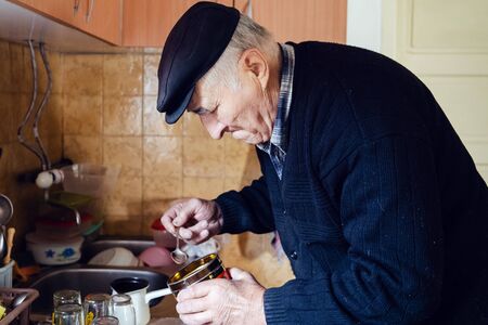 Senior man grandfather old pensioner farmer wearing black sweater and hat making a cup of coffee or tea cooking in the pot at home putting coffee to the pot with spoonの写真素材