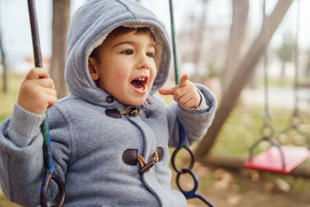 Portrait of a small little cute caucasian boy three years old having fun smiling on the swing in the park in winter or autumn day wearing coat pointing fingerの写真素材