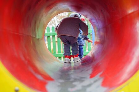 Small caucasian boy back view in the park on the playground in the red tunnel tube wearing winter coat in autumn day having fun playing walkingの写真素材